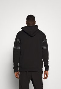 Svart huvtröja med dragsko i huvan, långa ärmar och horisontella loggor i svart och grått på ärmarna.