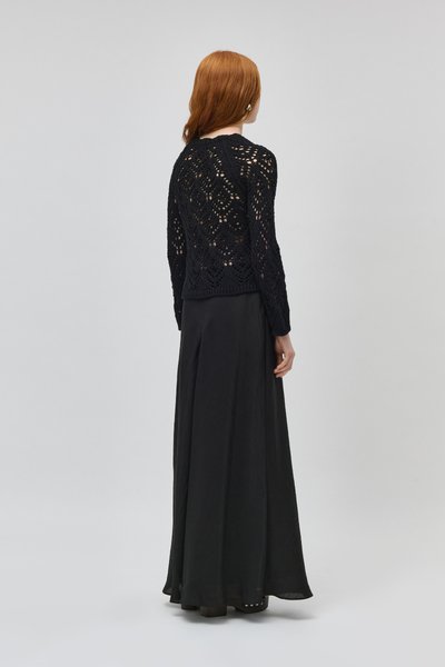 Femme aux cheveux roux portant un haut noir à manches longues en crochet et une jupe longue noire fluide, debout de dos sur un fond uni.