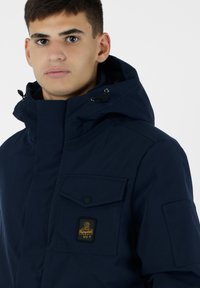 Giacca isolante blu navy con colletto alto, tasche frontali e patch con logo. Presenta un cappuccio regolabile e chiusure sicure.