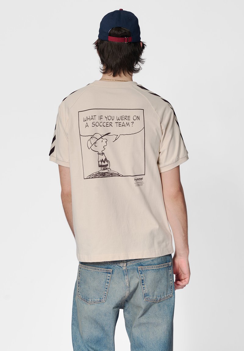 Beige katoenen T-shirt met een zwarte cartoonafbeelding op de achterkant met tekst. Het shirt heeft korte mouwen en contrasterende zwarte strepen aan de zijkanten.