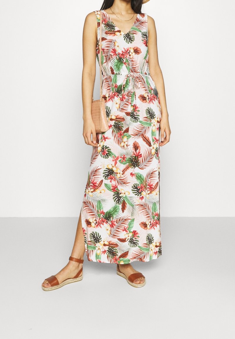 Femme portant une robe sans manches, longue jusqu'aux chevilles, à fleurs avec des feuilles et fleurs tropicales, des sandales marron et un sac bandoulière beige.