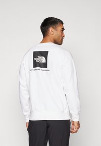 Witte sweatshirt met geribbelde manchetten en onderrand, voorzien van een grote zwarte rechthoekige grafiek op de achterkant met de merknaam en slogan.