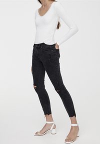 Jeans skinny negros rasgados con dobladillos deshilachados, combinados con una blusa blanca de manga larga que presenta un escote con ondas y detalles brillantes.