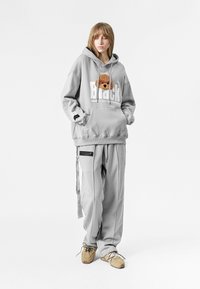 Sudadera gris con un gráfico de un oso y el texto "Black", combinada con pantalones de chándal grises a juego que tienen bolsillos con cremallera. Llevado con zapatillas beige.