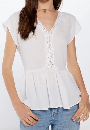Mujer vistiendo una blusa blanca de manga corta con detalle de encaje en el escote en V y bajo peplum, combinada con vaqueros denim azul claro y un collar con colgante dorado.