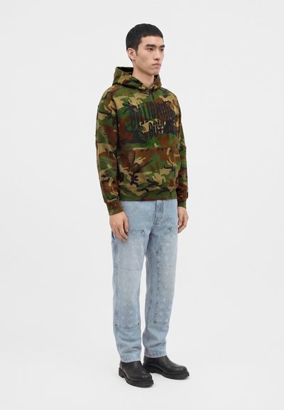 Young man standing wearing a green camouflage hoodie with text, light blue jeans, and black boots against a plain white background.