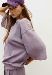 Przesadny, krótki sweter z rękawami w jasnofioletowym kolorze, wykonany z bawełny, z okrągłym dekoltem i miękką teksturą. Akcentowany fioletową czapką.
