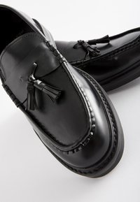 Mocassins en cuir noir avec des accents à pampilles, texture lisse, bout arrondi, et détails de couture complexes le long du bord.