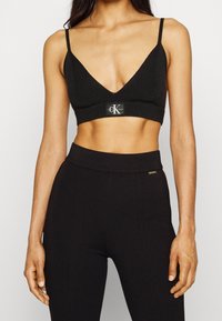 Musta bralette V-aukolla, teksturoidulla kankaalla, ohuilla hihnoilla ja brändilogo-patchilla vyötäröllä, yhdistettynä mustiin korkeavyötäröisiin housuihin.