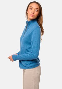Sweater zippé bleu en matériau texturé, avec un col haut et des manches ajustées. Porté par-dessus un pantalon clair.