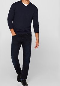 Pull à col en V bleu marine, jean foncé et baskets noires. Le pull a des manches longues et des poignets côtelés. Tissu lisse avec une légère élasticité.