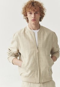 Beige bomberjakke lavet af tekstureret stof, med lynlås fra fronten, ribbede manchetter og sidelommer. Velegnet til afslappet brug.