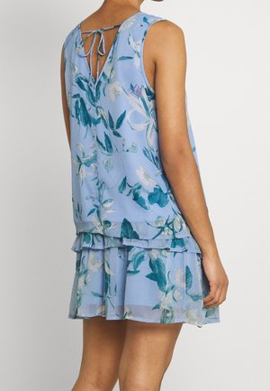 Mujer con un vestido sin mangas azul claro con estampado floral, bajo con volantes y detalle en la espalda en forma de V atada, vista desde atrás.