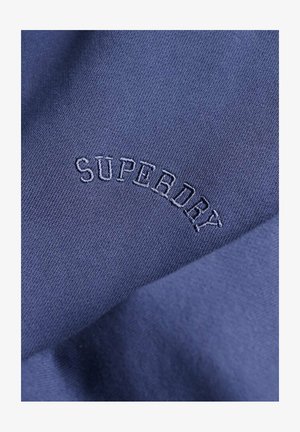 Tejido azul marino con una textura suave, que presenta la palabra "SUPERDRY" bordada en un diseño curvado.