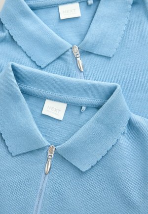Polo-uri ușor albastre dintr-un material tricotat texturat, cu un fermoar în față, guler ribbed și un etichetă cusută care indică "NEXT."