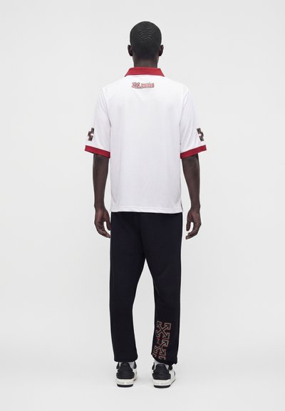 Personne vue de dos portant une chemise blanche à manches courtes avec un liseré rouge et un pantalon noir avec des motifs graphiques rouges sur la partie inférieure de la jambe.