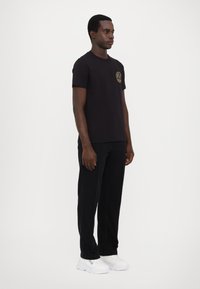 Tricou negru din bumbac cu guler rotund și logo circular auriu pe piept, asortat cu pantaloni negri și adidași albi.