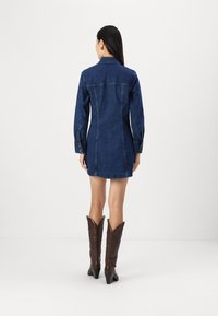 Jeans obleka z dolgimi rokavi, prilegajočega kroja, temno modre barve in rahlo obrabljenim robom. Na hrbtni strani ima šiv v obliki jermena in zapenjalne manšete.