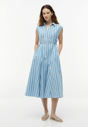Femme portant une robe midi sans manches à rayures verticales bleu et blanc, boutonnée, avec un col et des sandales, debout les mains dans les poches.