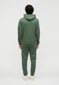 Zelená mikina s kapucňou a rebrovanými manžetami, doplnená o zodpovedajúce tepláky. Mäkká látka s voľným strihom a detailom vrecka na zadnej strane.