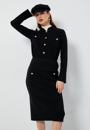 Ensemble noir deux pièces comprenant un haut à boutons avec des accents dorés, des manches longues, et une jupe crayon avec poches avant et boutons dorés.