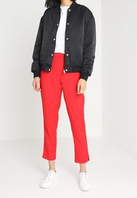 Schwarze Satin-Bomberjacke mit Druckknöpfen, kombiniert mit leuchtend roten, geraden Hosen. Weiße Sneakers runden das Outfit ab.