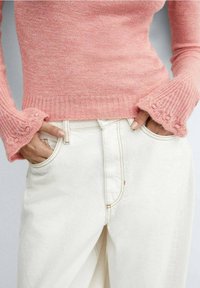 Hands in front pockets of off-white jeans, wearing a fitted pink knit sweater with flared sleeves and textured cuffs.