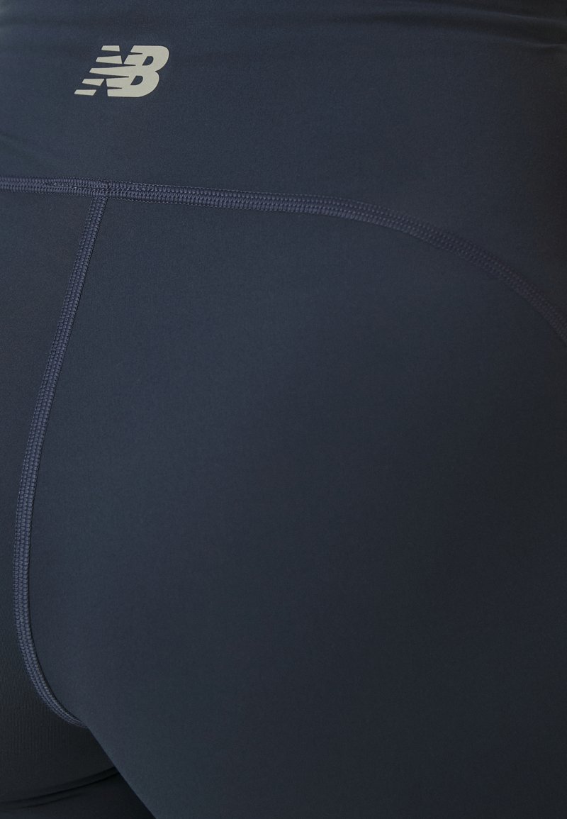 Gros plan sur des leggings de sport bleu marine avec des lignes de couture visibles et un petit logo New Balance blanc près de la taille.