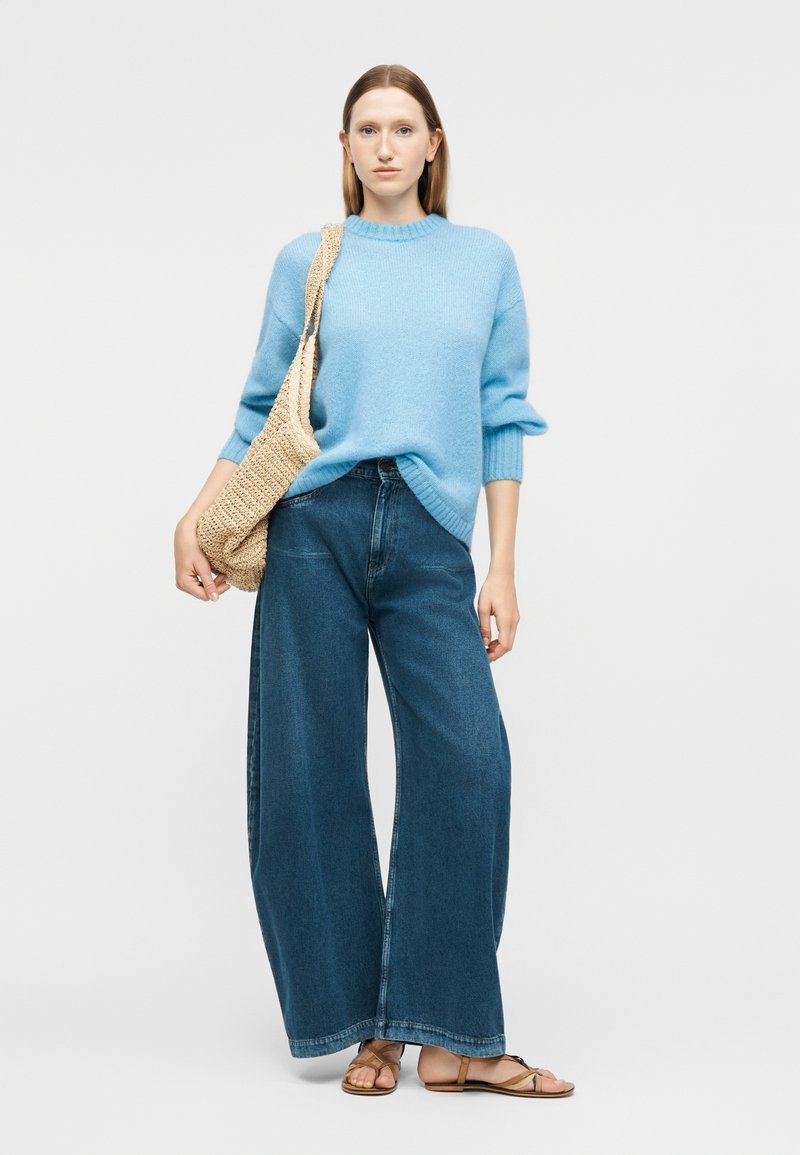 Frau trägt einen hellblauen Pullover, dunkelblaue Jeans mit weitem Bein, braune Sandalen und eine beigefarbene, gewebte Umhängetasche.