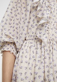 Robe fleurie en tissu beige texturé avec des motifs floraux violet clair et noirs, manches à volants et taille froncée.