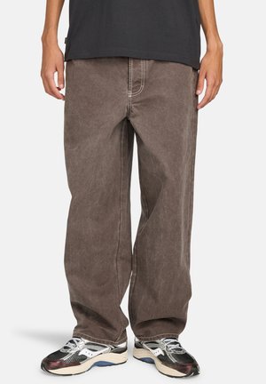 Brown loose-fit denim pants with visible stitching and a faded texture, paired with black and silver sneakers featuring mesh accents.