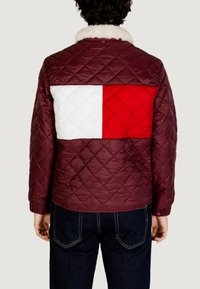Giacca trapuntata color bordeaux con pannello bianco e rosso sulla schiena, caratterizzata da un colletto in shearling e polsini elasticizzati. Indossata con jeans di denim scuro.