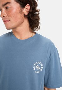 Mladý muž se středně dlouhými hnědými vlasy, oblečený v modrém tričku Quiksilver 1969, dívající se doleva na jednobarevném pozadí.