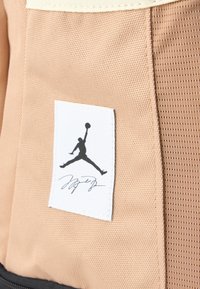 Dzeltenīgi brūns audums ar baltu birku, uz kuras ir melns Jumpman logo un paraksts, parasti sastopams sporta somās vai aprīkojumā.