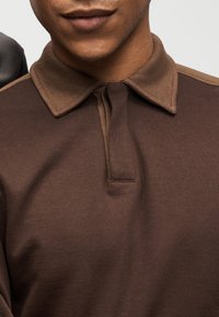 Καφέ πόλο πουκάμισο με υφασμάτινη υφή, κλασικό γιακά και μπροστινή πλακέτα με κουμπί. Ο σχεδιασμός περιλαμβάνει αντιθετικές λεπτομέρειες στους ώμους.