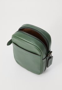 Borsa a tracolla verde in pelle con una superficie liscia, bordi arrotondati e doppi zip. Presenta un'etichetta laterale e un design semplice e pulito.