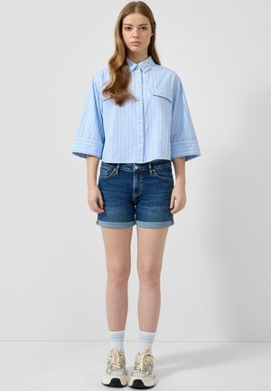 Jeune femme debout portant une chemise à rayures bleu clair boutonnée, un short en jean retroussé, des chaussettes blanches et des baskets beige sur un fond gris uni.