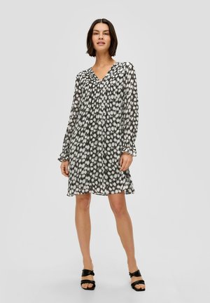 Robe à pois noir et blanc, en tissu léger, décolleté en V, manches longues avec poignets froncés, silhouette fluide, longueur genoux, associée à des sandales noires.