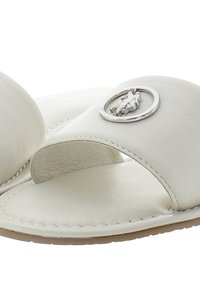 White leather slide sandals featuring a wide band, open toe, and a circular metallic emblem with detailing on the strap.