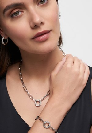 Silberne Kettenhalskette und Armband mit runden Akzenten und funkelnden Verzierungen; das Modell trägt Creolen.