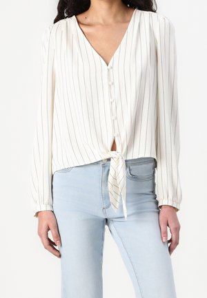 Femme portant un chemisier blanc à manches longues avec de fines rayures verticales noires, boutonné à l'avant, noué à la taille, associé à un jean bleu clair.