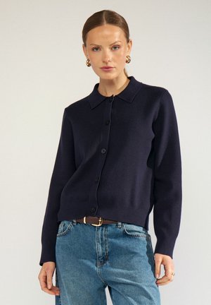 Femme portant un cardigan bleu marine foncé boutonné, un jean bleu, une ceinture marron et des boucles d'oreilles créoles en or sur un fond clair uni.