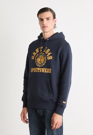 Mörkblå hoodie i bomullsblandning med framficka. Utrustad med en gul tryckt logotyp som säger "GANT 1949 SPORTSWEAR" och snodd i huvan.