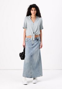 Light blue short-sleeved blouse with button details and tie front, paired with a long light denim skirt. White sneakers and a black patterned bag.