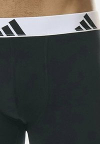 Acercamiento de leggings deportivos negros con una cinturilla blanca que presenta un logo parcial en negro de Adidas.