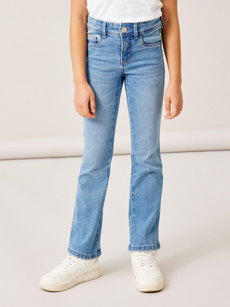 Jeans de mezclilla azul claro acampanados con un diseño clásico de cinco bolsillos y trabillas para el cinturón. Combinados con zapatillas blancas. Textura suave.