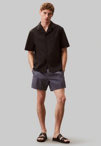 Camisa negra de manga corta con bolsillo, combinada con pantalones cortos grises y sandalias negras. Tejido suave con una textura sutil y un diseño simple.