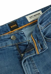 Modré denimové nohavice s strieborným zipsom a zapínaním na gombík. Štítky obsahujú nápisy "BOSS" a "SOFT MOTION." Interiér má hladkú tkaninovú podšívku.