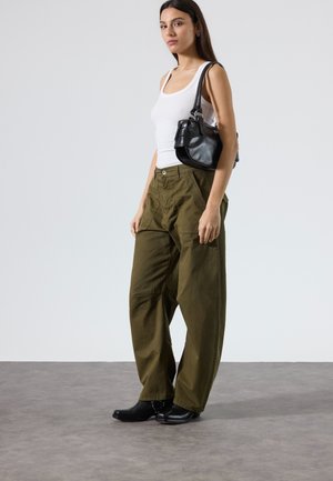 Frau steht, trägt ein weißes Tanktop, lockere olivgrüne Hose, schwarze Stiefel und trägt eine schwarze Schultertasche auf ihrer rechten Schulter.