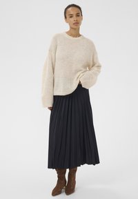 Bēša adīta džempera ar apaļu kakla izgriezumu un pazeminātām pleciem, kombinēta ar tumši zila, piegriezuma līdz zemes, krokojumu svārkiem un brūnām potīšu zābaku.